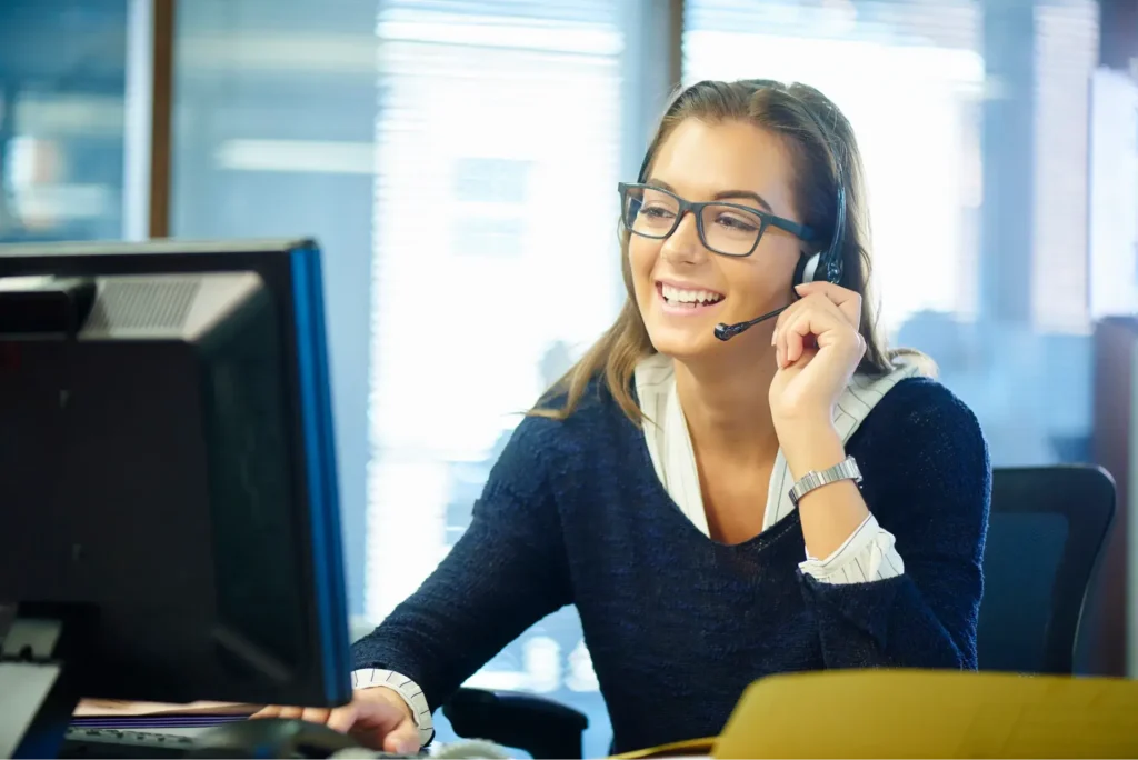 Photo of a smiling woman, sitting at a desk working on a computer while talking a phone.